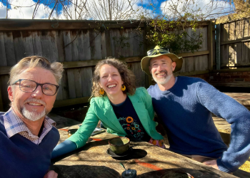 A man with a beard smiling at the camera, opposite him, also smiling at the camera from the other side of a table is a woman in a green jacket and a man close and next to her wearing a hat.